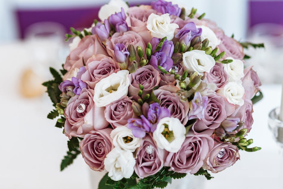 A close-up view of a floral bouquet featuring soft pink roses, white lisianthus, and purple freesia, arranged in a rounded, balanced style. The flowers appear fresh with delicate petals and vibrant co
