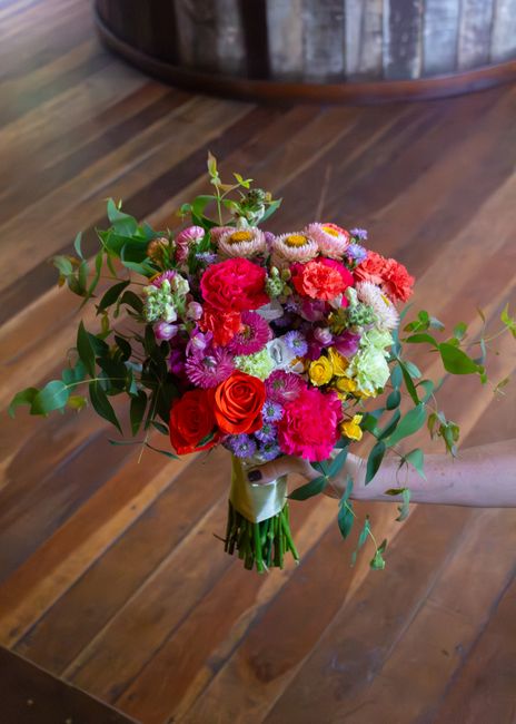 A vibrant bouquet of fresh flowers held by a person's hand, featuring a mix of pink, red, purple, white, and yellow blooms. The arrangement includes roses, daisies, and carnations, complemented by lus