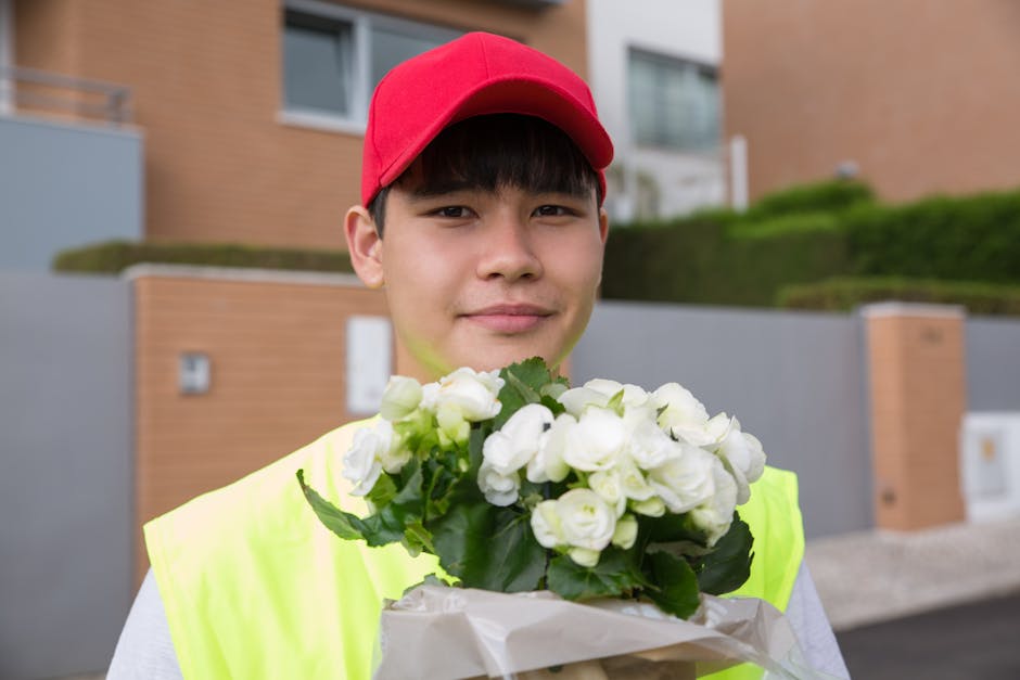 A young man wearing a red cap and a fluorescent yellow vest holds a bouquet of fresh white flowers, possibly roses or similar blooms, wrapped in white paper. He is standing outdoors in front of modern