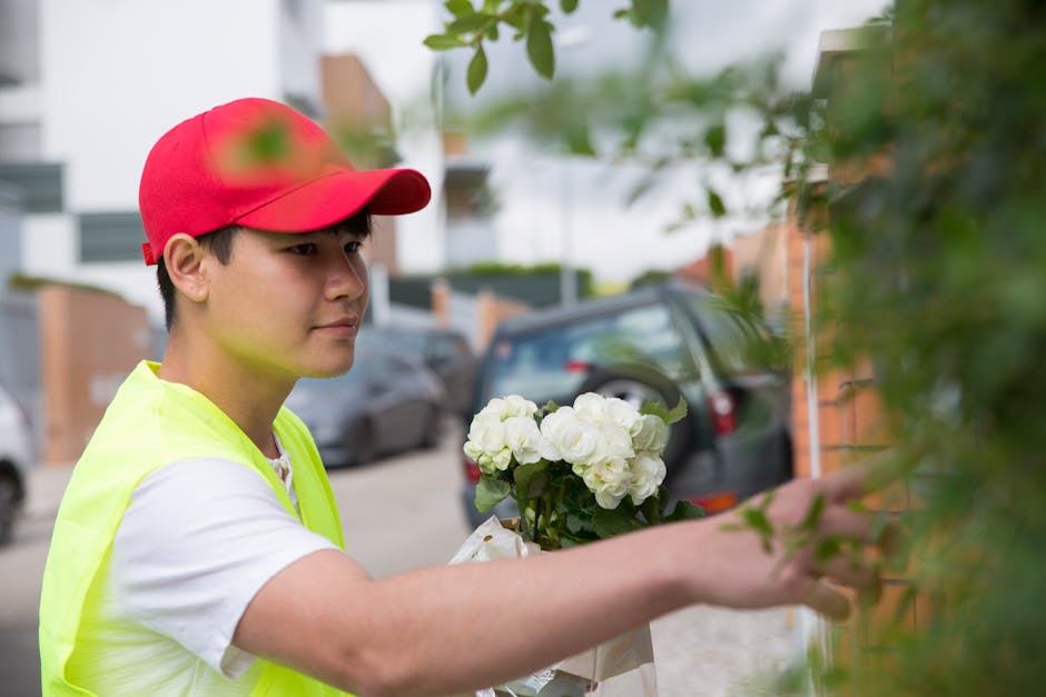 A delivery person wearing a red cap and a high-visibility yellow vest is seen placing a bouquet of fresh white hydrangeas wrapped in white paper against a green leafy shrub outside a residential build