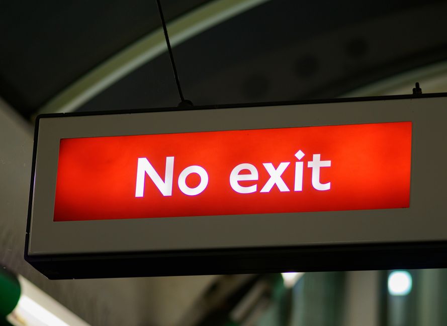 A rectangular illuminated sign hanging from a ceiling, displaying the words 'No exit' in bold white letters on a bright red background. The sign is suspended by a black wire, with the background showi