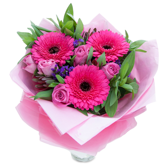 There is a playful grace to this bouquet: three wide-eyed Gerbera daisies in a confident fuchsia hue burst forward, their many petals arranged like a joyful fan around darker, almost black centres that give depth and contrast. Nestled among them, lavender-pink roses soften the composition-each rose is a study in tender curvature with petals that cup inward, suggesting newness and careful handling. Tiny sprays of purple statice are tucked into the negative spaces, adding flecks of cool colour and a papery texture that recalls wild hedgerows beyond Epping Forest. Supportive foliage-broad, lush leaves reminiscent of salal or eucalyptus-wraps the flowers in cool, verdant tones and imparts a faint, green, slightly resinous scent when you lean in. The florist's touch is evident in the finishing: an inner sheet of pale pink, matte tissue paper creates a gentle base while an outer layer of translucent fuchsia-pink cellophane, slightly crinkled, adds a glossy whisper and ties the palette together. The lower edge reveals a clear glass vase rim, speaking to the bouquet's readiness to be placed in water and enjoyed immediately. Soft, even lighting in the image accentuates the subtle differences between hot fuchsia and dusty rose, while the composition feels intimate-as if meant to be handed over on Epping High Street or left as a surprise on a doorstep after a visit to nearby Loughton. It's a thoughtful arrangement for celebrations small and large, a visual hug that brings cheer and considered craftsmanship.
