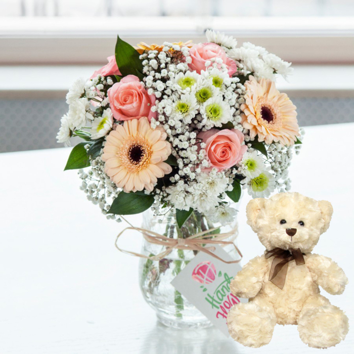 On a gleaming white surface that captures a whisper of reflection, a romantic anniversary gift set is arranged with quiet elegance, reminiscent of the tranquil light that filters through homes around Epping Forest. The centre of the scene is a clear glass jar vase, simple yet charming, filled with a compact bouquet of pastel flowers. Blush pink roses, gently unfolding, lean into peach gerbera daisies whose velvety petals surround distinctive dark brown centres. These focal blooms are softened by drifts of tiny white baby's breath, threading delicately throughout the arrangement, and by rounded heads of small white chrysanthemums with vivid lime-green centres that add a touch of freshness and vitality. The stems inside the vase form a lattice of greens and soft browns, grounding the airy colours above. Encircling the vase neck is a band of natural twine, tied in an unfussy bow, from which dangles a small white rectangular tag. The tag bears a simple pink heart and flowing green script, suggesting a handcrafted note or locally made gift, like something curated for a couple celebrating in Epping. To the right, a cream teddy bear sits upright, its body turned slightly towards the bouquet. Its fur appears thick and plush, with a touchable, cosy texture. Dark bead eyes, a modest brown nose, and a neatly tied dark brown satin ribbon at the neck give the bear a classic, timeless charm. Behind them, the background is softly washed in light and blur, ensuring that the eye rests on this affectionate duo - a vase of gentle blooms and a cuddly bear, together capturing the warmth of love, comfort, and shared memories.