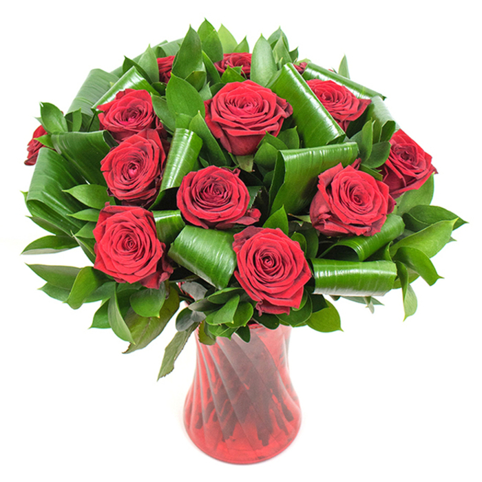 This image showcases a professionally composed bouquet of roughly a dozen deep crimson roses, each blossom at the peak of its bloom with dense, velvety petals spiralling into a sumptuous centre. The rich red is intensified by even studio lighting, which picks out the soft texture of the petals and the subtle shadowing between layers. Surrounding the roses is a generous, sculptural mass of dark green foliage that provides both contrast and support: narrower glossy leaves form a verdant backdrop while larger, lustrous leaves are artfully rolled and curved into dramatic loops and tubes, creating an architectural counterpoint to the softness of the roses. The floral composition rests in a tall, slender ruby glass vase with a faint vertical ribbing that catches light and accords a graceful sense of height and stability; the vase's narrow waist and flared top frame the bouquet like a jewel. Taken together, the arrangement balances classic romantic symbolism with contemporary floristry techniques - careful leaf folding, measured negative space, and a lustrous finish. You can almost sense the cool, smooth weight of the glass in your hands and the gentle, familiar fragrance of red roses, a scent that carries memory as much as beauty. Ideal for a romantic dinner on Bell Common, a thoughtful anniversary surprise for a neighbour near Coopersale, or as a luxurious centrepiece for an intimate gathering, this bouquet reads as an expression of enduring love and considered design, rooted in the quiet character of Epping and its leafy surroundings.