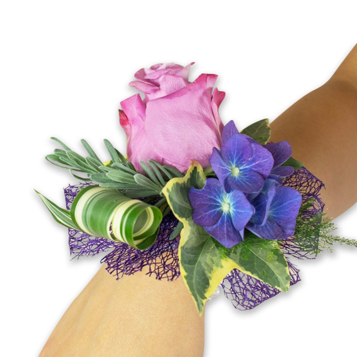 Displayed diagonally across a pale forearm against a pristine white background, this wrist corsage is an expression of refined celebration crafted with attention to texture and tone. At its centre a full, softly unfurling pink-mauve rose offers a romantic heart, its velvety petals showing a gentle gradient from a lighter center to a slightly deeper magenta at the ruffled edges. Surrounding the rose are three prominent clusters of deep indigo-purple hydrangea; each tiny floret carries a subtle blue-white core that adds sparkle and visual depth. The arrangement is grounded with glossy variegated ivy, the dark green leaves kissed with creamy margins, and with slender, silvery-green foliage-suggestive of eucalyptus-woven behind the blooms to create a soft, scented backdrop. A contemporary flourish comes from a broad green leaf striped in white, artfully curled into a tight spiral to introduce sculptural movement. Below, a layer of open, dark purple mesh peeks through, amplifying the hydrangeas' tones and providing a lacy base. As a local florist I think of how this piece would complement an understated dress for a wedding at St. John's Church or a prom near Epping Station; Flower Delivery Epping prepares pieces like this to feel both personal and memorable. The corsage evokes cool summer air, a delicate herbal scent, and the quiet pleasure of wearing a handmade floral accent-an elegant, tactile keepsake for special moments in and around Epping.