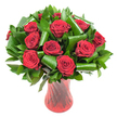 A sumptuous, rounded bouquet of a dozen or more deep crimson roses sits poised in a tall, translucent ruby-red glass vase, captured against a clean white backdrop so every velvety petal and curled leaf reads clearly. Each rose is fully open with tightly spiralled centres that ease into soft, satiny outer petals; the blooms are so richly pigmented they read almost wine-dark at the heart, mellowing to a vibrant crimson at the edges. Surrounding the rose heads is an abundant collar of glossy, dark green foliage - dense Ruscus- or Salal-like leaves create volume and a verdant base while broader emerald leaves, reminiscent of Aspidistra, have been expertly folded and rolled into sculptural loops and spirals that frame and lift each bloom. The leaf folds add an architectural, modern touch against the classic romance of the roses, catching the soft, even studio light to reveal smooth, reflective textures. The vase is a slender, slightly flared ruby glass with a subtle ribbed twist that runs vertically, throwing thin highlights down the curve and anchoring the composition with elegant lines. Together the elements show a careful hand: balanced proportion, thoughtful negative space, and a tactile contrast between velvety petals and glossy greens. The arrangement smells faintly of classic rose perfume - warm, gentle, and familiar - and radiates an air of refined passion, perfect for anniversaries, a Valentine's centrepiece, or a meaningful gift. A local florist in Epping might recommend this arrangement for a romantic table at a cottage on the High Street or a surprise delivered to a home near Epping Forest, where the deep reds echo autumnal woodlands nearby.