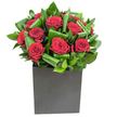 In this elegantly composed arrangement, rich crimson roses form the immediate focal point, each bloom unfolding in velvety spirals that catch the eye with a soft, luxurious depth. The flowers are grouped tightly to read as a single, emotive statement-romantic, assured, and quietly dramatic-while broad, glossy green leaves are curled and layered around the cluster to create a sculptural rhythm. Those leaves lend movement and contrast: smooth, almost waxy surfaces that reflect the clean, modern light in bright highlights and deepening shadows, giving the bouquet a sense of three-dimensional clarity. The roses sit in a matte black square vase that grounds the composition with an urban, minimalist edge; its blocky silhouette offsets the organic curves above and makes the crimson tones sing. The lighting is crisp and contemporary, as if photographed near a large studio window, revealing subtle textures in each petal and the faint sheen of morning dew. A gentle fragrance seems to lift from the arrangement-classic rose softened by green, almost tropical leaf notes-inviting a slow, appreciative breath. As a florist who spends mornings checking stems and placing final touches in Epping High Street, I imagine this design as suited to rooftop dinners, gallery openings, or simple declarations of affection. It reads as both a statement of passion and an object of calm refinement, perfect for anniversaries, heartfelt apologies, or to bring a polished, living warmth into a modern home on the edge of Epping Forest.